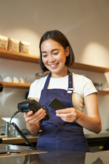 Portrait of smiling asian barista, coffee shop employee using POS terminal and credit card, helps...