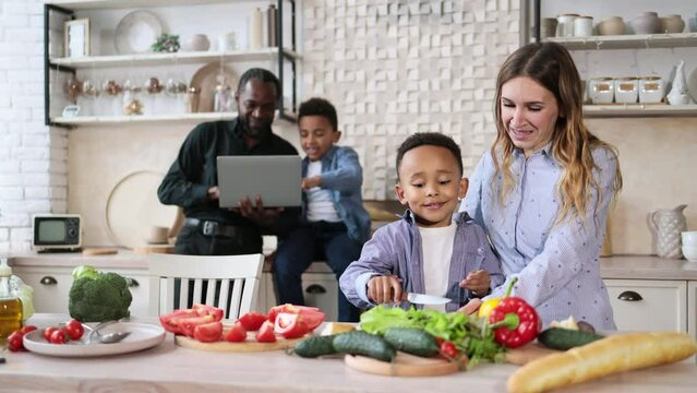 Pretty mother and son preparing tasty food at kitchen. Mommy teaching lovely kid to cook while happy father and child boy spending time using laptop computer. Healthy meal and dinner preparation.
