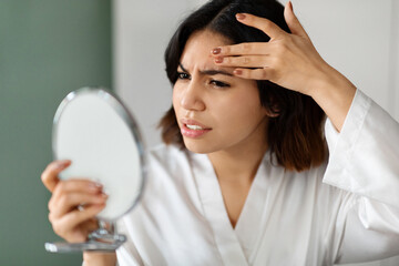 Upset arabic woman looking at hand mirror, checking skin