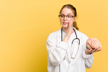 Young doctor caucasian woman holding a brain isolated on yellow background is saying a secret hot...