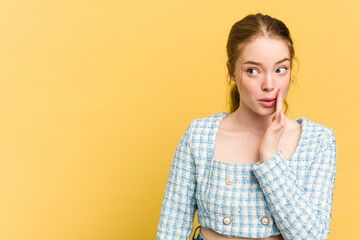 Young caucasian redhead woman isolated on yellow background is saying a secret hot braking news and looking aside