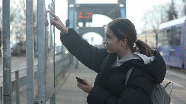 teenage girl with smartphone in hand stands next to the Wroclaw public transport schedule boar