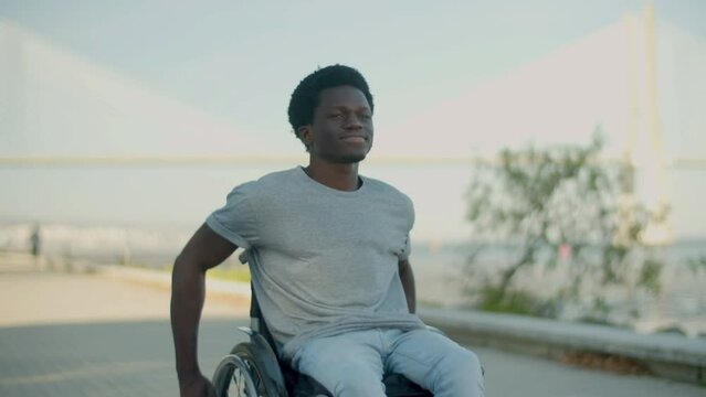 Young Black man riding in wheelchair along seashore on warm summer day. Happy guy with paraplegia spending time at seafront. Front view. Disability, leisure time, lifestyle concept. 