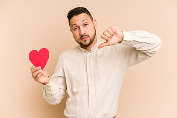 Adult latin man holding a red heart isolated showing a dislike gesture, thumbs down. Disagreement...