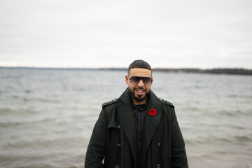 Bi racial model standing on board walk with water behind him winter time with sunglasses and green jacket on 