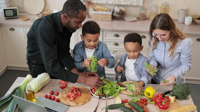 Multinational family cooking healthy salad on light kitchen in weekend. Cute boys helping their young parents tearing freshly washed, fresh clean leaves of lettuce into bowl, preparing healthy salad.