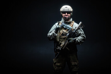 soldier in military equipment with a rifle smiles at night, a commando in uniform against a black...