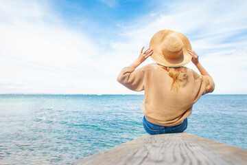 girl in a sun hat sits on a pier against the background of the sea and looks at the horizon