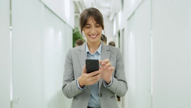 Portrait of successful young female worker of company walking in corridor scrolling using smartphone greeting colleagues. Attractive stylish woman employee wearing smart style clothes in office.