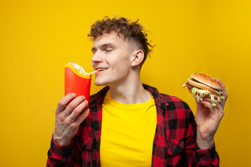 curly guy eats french fries and holds a burger on a yellow background, a satisfied man eats fast...