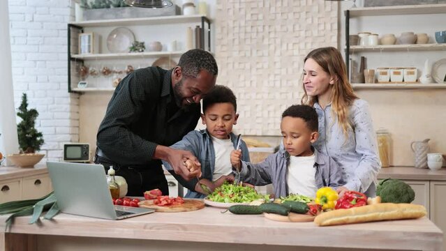 Young happy mom, dad and little sons adding olive oil to the salad while cooking in kitchen together. Happy multiracial parents and small boys kids have fun preparing healthy food at home.