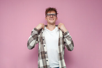 confident guy in glasses puts on a plaid shirt on a pink background