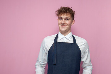young waiter in white shirt and apron smiling on pink background, portrait curly guy barista worker...