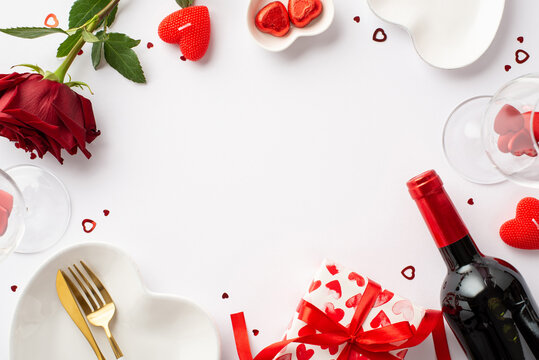St Valentine's Day concept. Top view photo of heart shaped tableware giftbox cutlery candies wine bottle glasses candles rose and confetti on isolated white background with blank space in the middle