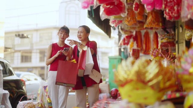 Asian mother and daughter using navigator on mobile phone while shopping home decorative ornaments and joss paper for celebrating Chinese Lunar New Year festival together at Chinatown street market.

