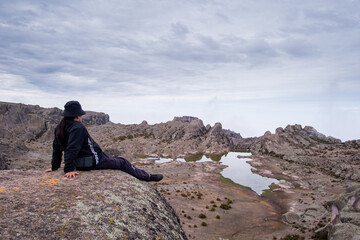 half-sitting latin man with his arms supporting her, with a lagoon and a valley of rocks in the background.