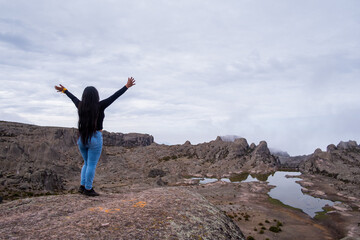 Latin woman with her back turned and arms raised, standing on the edge of a cliff with a lagoon in the background.