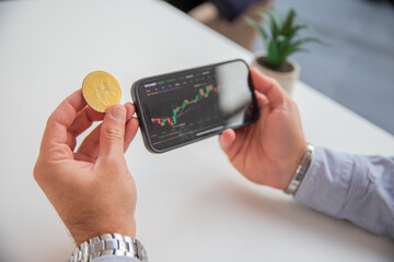 A trader is holding a bitcoin coin and analyzing the market charts on his smartphone.