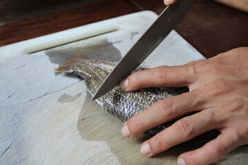 An asian man is cutting a fish with a knife on a white cutting board in the kitchen