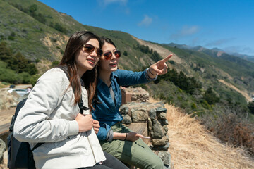 asian Korean girl showing friend a tourist attraction in space with pointing gestures as they are sitting enjoying landscape in nature at hillside in California usa