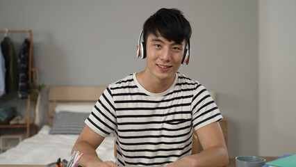 asian college Japanese boy learning from home with a headset is looking at camera and talking with ok hand gesture during a virtual online course in the bedroom at home