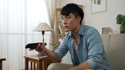 impatient asian Japanese guy shaking head and feeling dissatisfied while channel surfing with a...