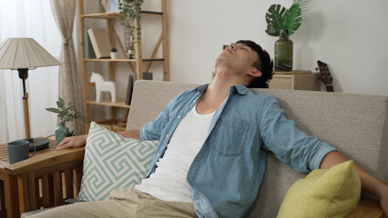 bored asian male turning off tv with controller and leaning back on couch with spreading arms feeling depressed while staying at home during quarantine