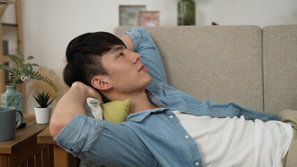 sleepy asian male reclining on sofa with a yawn is staring into space after putting down his phone...