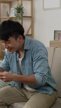 Vertical Screen: overjoyed asian chinese male checking number on sofa is waving clenched fists with excitement and throwing lottery tickets in air while cheering for winning at home