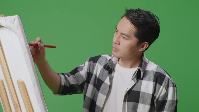 Close Up Of Asian Male Artist Shaking His Head Unsatisfied With The Picture On Canvas While Painting By Oil Paints And Brush In The Green Screen Studio
