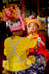 Vertical view of little Asian boy wear tradditional cloth of young novice buddhist monk help to put on makeup of his coworker with concentrate and happy.