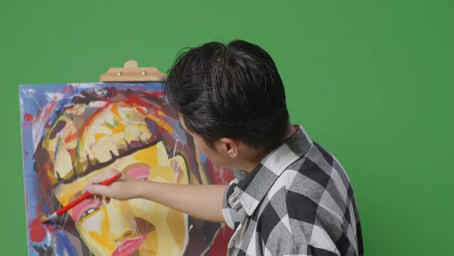 Close Up Back View Of Asian Male Artist Enjoys Painting A Girl On Canvas By Oil Paints And Brush In The Green Screen Studio
