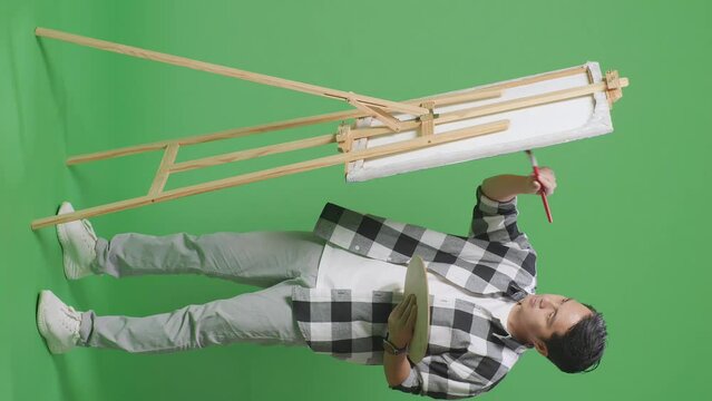 Full Body Of Asian Male Artist Smiling To Camera While Painting On Canvas By Oil Paints And Brush In The Green Screen Studio
