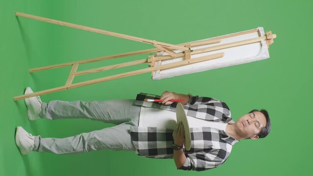 Full Body Of Asian Male Artist Shaking His Head Then Having A Headache While Painting On Canvas By Oil Paints And Brush In The Green Screen Studio
