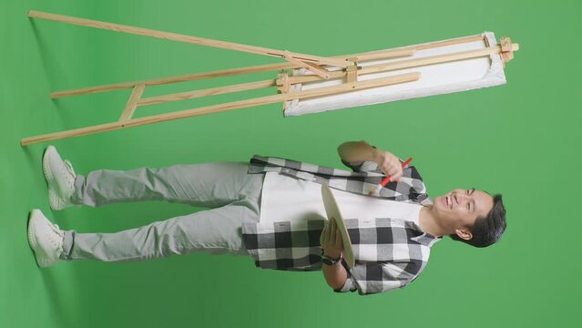 Full Body Of Asian Male Artist Celebrating Finish Painting On Canvas By Oil Paints And Brush In The Green Screen Studio

