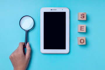 Hand holding magnifying glass with tablet smartphone and wooden cube lettering SEO on blue background.  Search Engine Optimization concept.