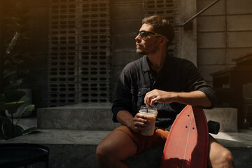 attractive man in a black shirt and glasses sits in a stylish cafe interior with his skateboard and drinks a cold caffeinated drink.