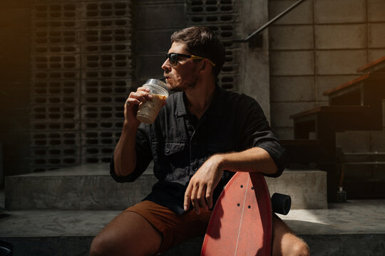 Modern Man in a black shirt and glasses sits in a cafe with his skateboard and drinks a cold drink.