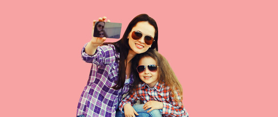 Portrait of happy smiling mother and child taking selfie by smartphone together on pink background