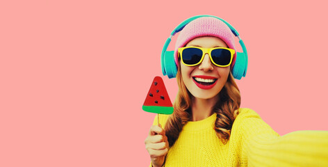 Summer fresh colorful portrait of laughing woman model taking selfie in headphones listening to music with fruit juicy lollipop or ice cream shaped slice of watermelon on pink background