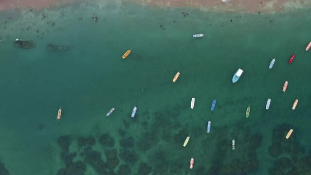 Left trucking bird's eye top-down aerial drone shot over crystal clear green water with anchored sailboats, rocks, and seaweed below in Porto de Galinhas or Chicken Port beach in Pernambuco, Brazil.