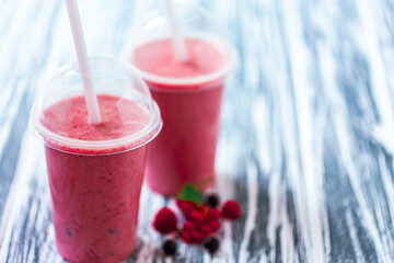 Front view of two berries milkshakes in plastic cups with ingredients on wood table. Space for text