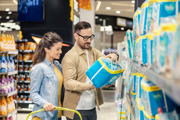 A young parents are standing at supermarket and purchasing diapers for their baby.