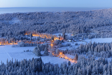 Amazing winter landscape scene during pleasant sunrise. Snow covered trees and meadows, city lights still on shining through orange. A view from hill down below. 