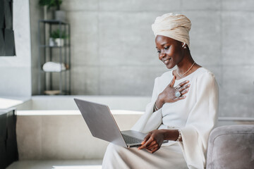 Grateful African woman in white turban and dress sitting at home makes video call using laptop...