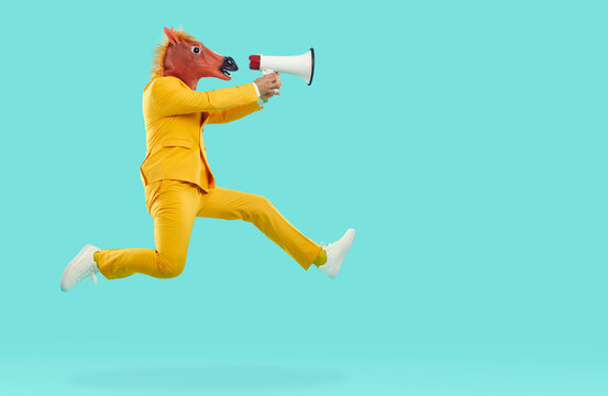 Man in horse head mask jumping and shouting in megaphone. Side view of eccentric guy wearing stylish yellow suit and animal mask cheering up, making advertisement with loudspeaker