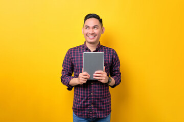 Smiling young Asian man holding digital tablet and looking aside at copy space isolated over yellow background