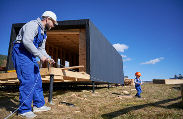 Father with toddler son building wooden frame house. Male builder and kid playing with tape measure on construction site, wearing helmet and blue overalls on sunny day. Carpentry and family concept.