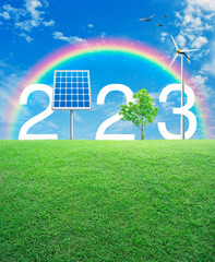 2023 white text with solar cell, wind turbine and growing tree on green grass field over rainbow, birds and blue sky with white clouds, Happy new year 2023 ecological cover concept