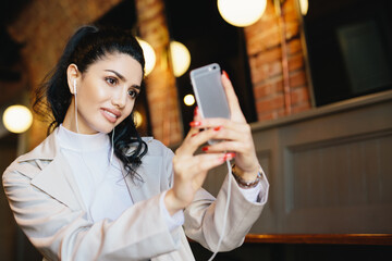 Beautiful woman with dark hair tied in pony tail making selfie while sitting in cafe using her...